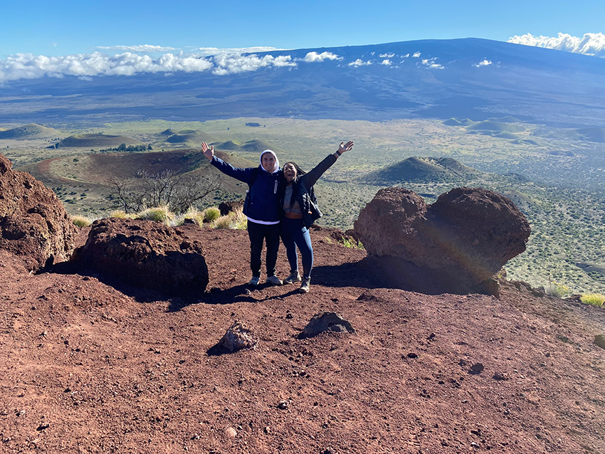 Andrew and Kym stand atop a mountain with their arms outstretched. They appear to be above the clouds, volcanic hills are visible in the distance. 